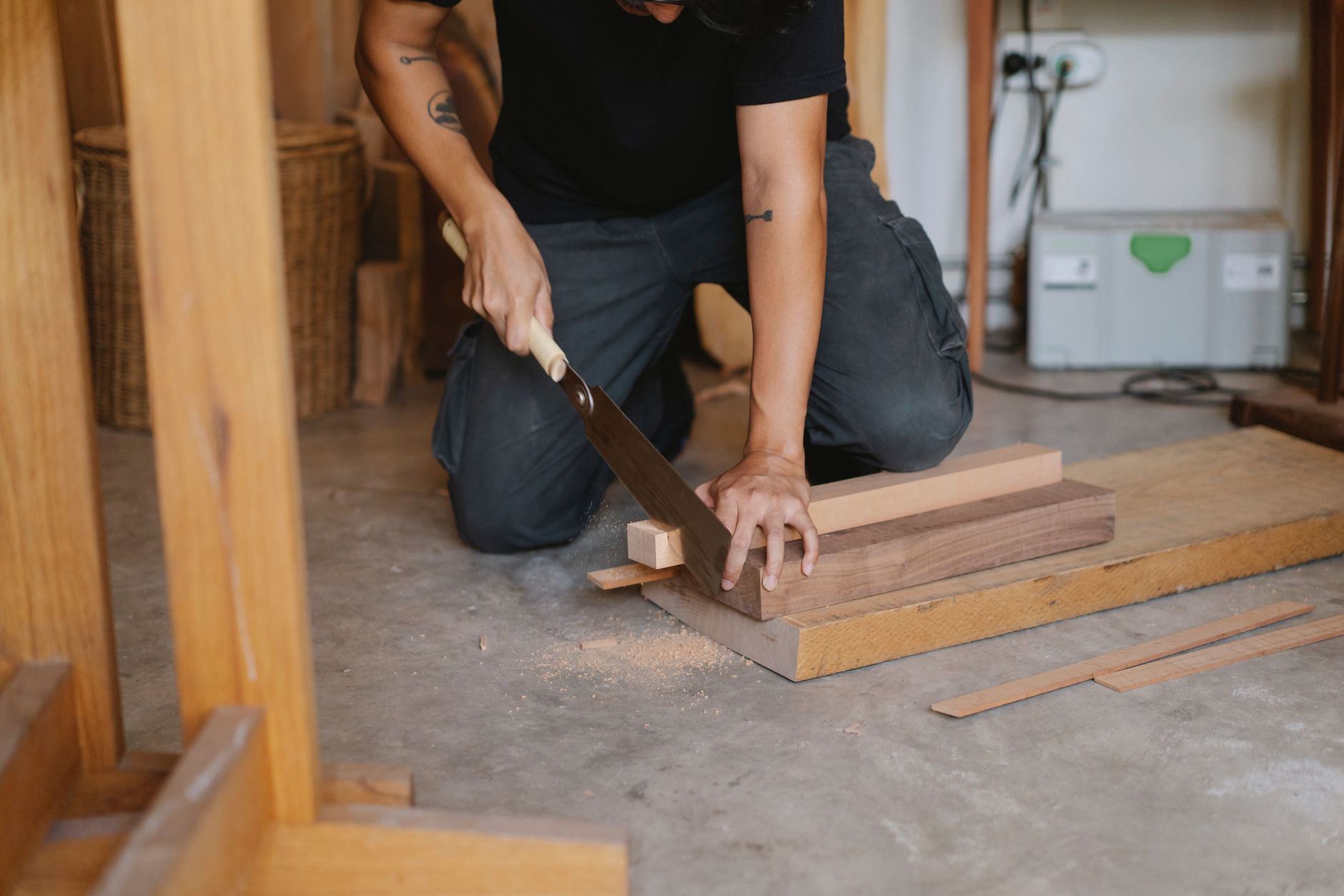 Unrecognizable skilled male carpenter with tattoo cutting wooden planks with hand saw while sitting on floor in professional studio during work
